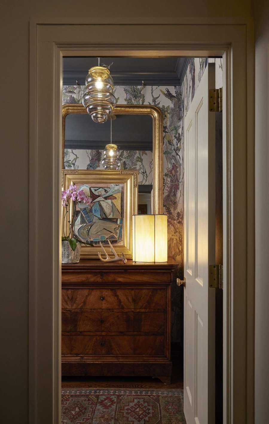 View through a doorway to a burl wood chest, gilt mirror, and glowing lamp against botanical wallpaper, designed by Kimberly Knight of Twig and Trove Interiors